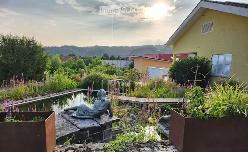 a garden with a pond, plants, flowers, statue, wooden walkway, house in the background, mountains
