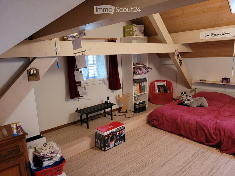 Cozy attic bedroom with slanted wooden ceilings, a large bed with red bedding, a red stool, a shelf, a bench, and various decorative items.