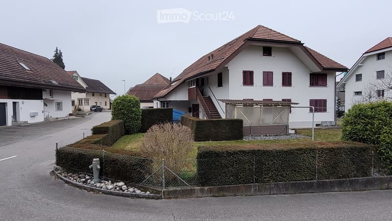 A two-story residential house with a red-tiled roof, white exterior walls, and multiple windows with shutters. The house features a staircase leading to an upper level and is surrounded by neatly trimmed hedges and a small garden area.