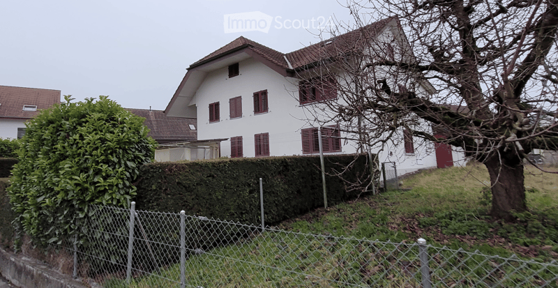 Two-story white house with red shutters and a brown tiled roof, partially obscured by a hedge and a tree.