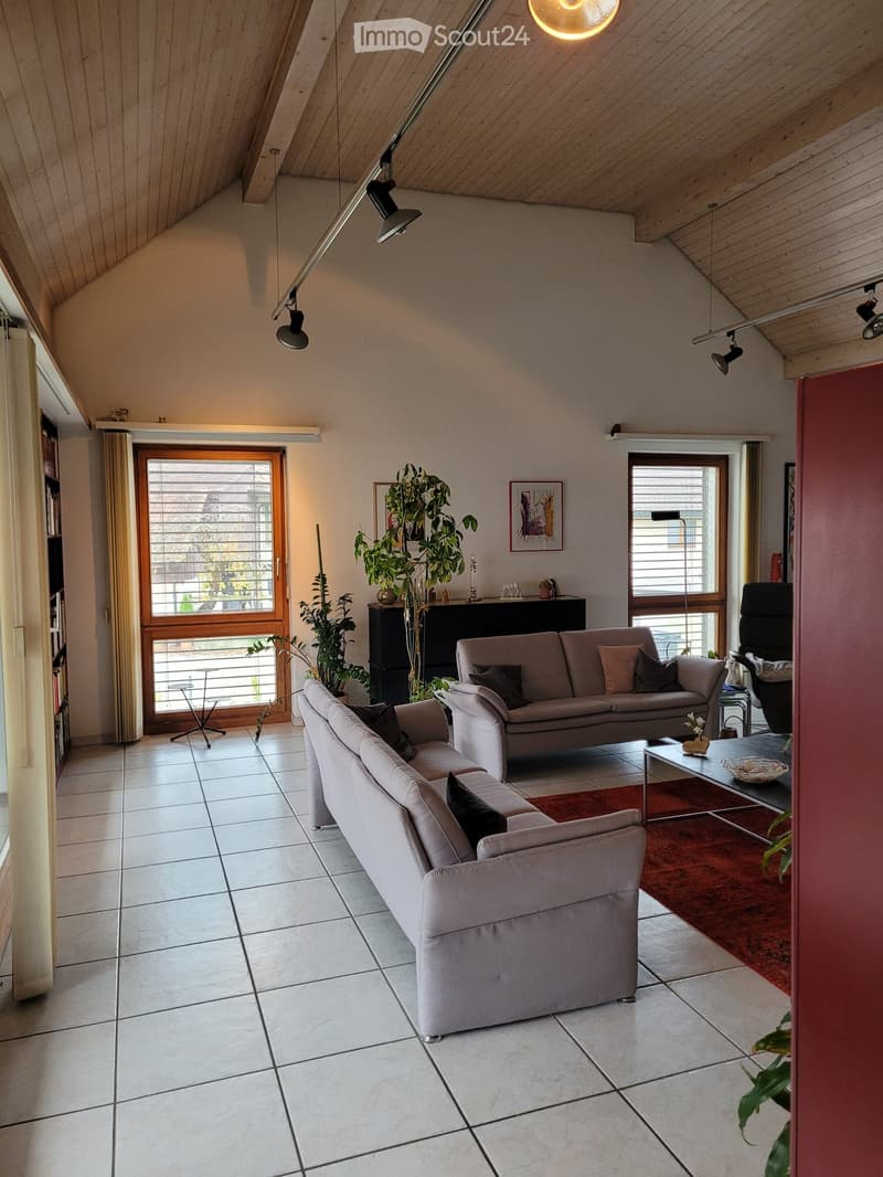Modern living room with two sofas, coffee table, side table, and large windows. Features a slanted wooden ceiling and pendant lights.