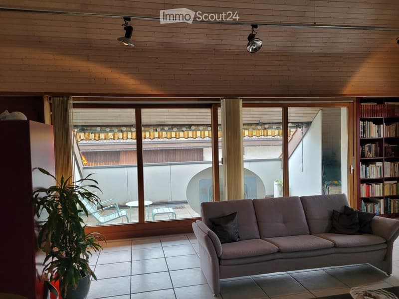 Bright living room with wooden ceiling, large windows opening to a balcony, sofa, bookshelf, potted plant, and outdoor seating visible through the windows.