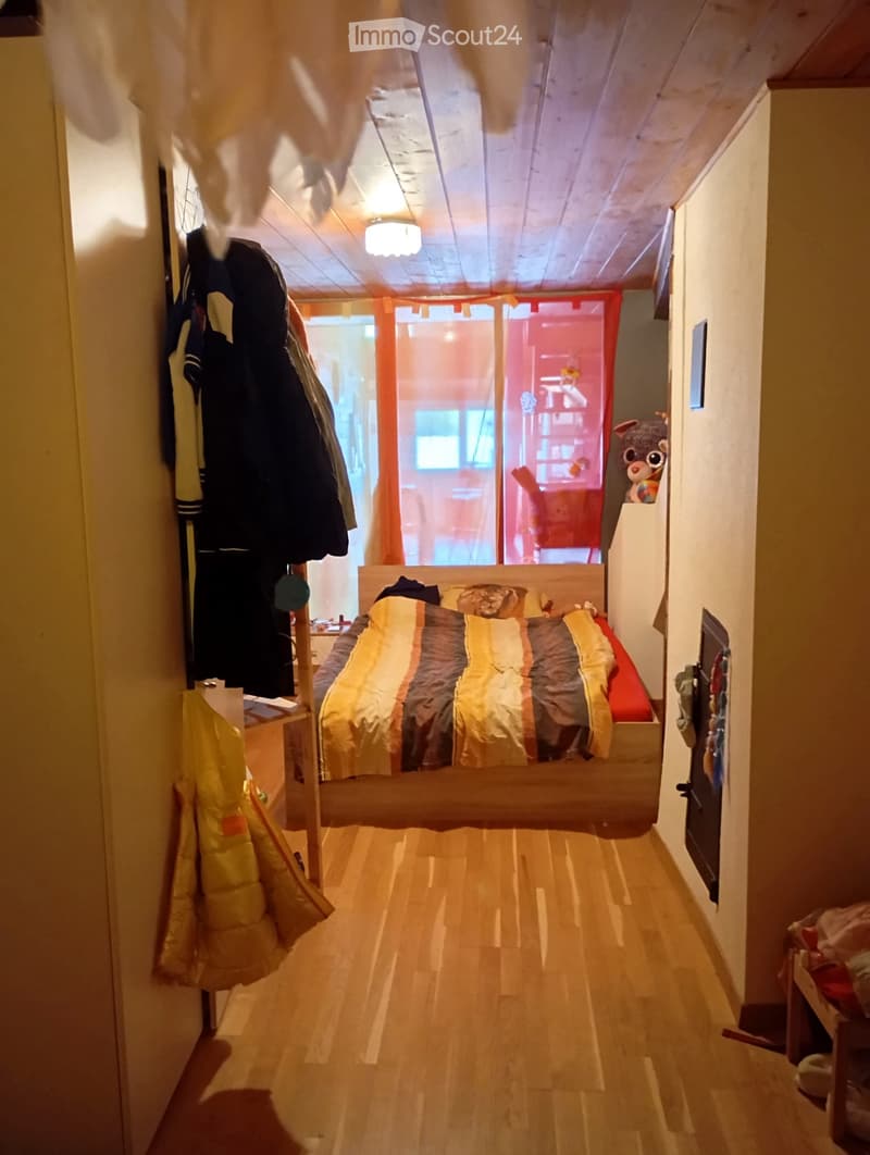 Bedroom with bed, wooden floor, ceiling lamp, clothes hanging on wall, window with curtains, fireplace, toys on shelf