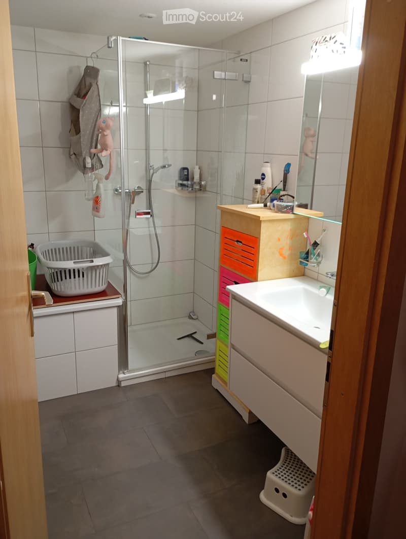 white tiled walls, glass shower enclosure, white sink, wooden cabinet, storage boxes, mirror, toothbrush holder, basket, green bucket