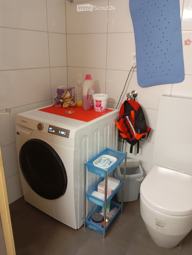 A small laundry room with a washing machine, a shelf, a trash bin, and a toilet.