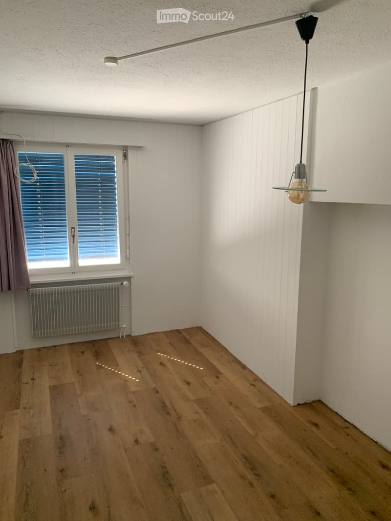 Empty room with wooden flooring, white walls, a window with blinds, a radiator, and a hanging light fixture.