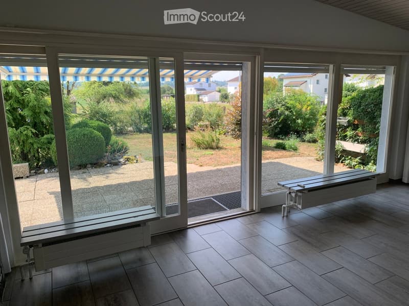 Empty room with large sliding glass doors opening to a garden, tiled floor, radiators, and a view of greenery and neighboring houses.