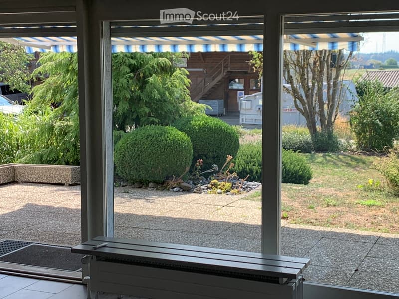 View from inside a room through large windows showing a well-maintained garden with shrubs, a small pond, and a wooden staircase leading to an outdoor structure.