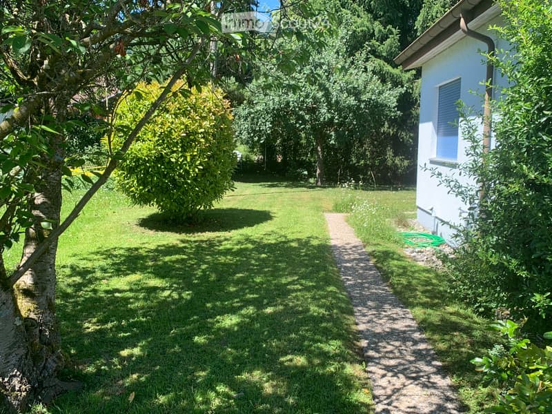 Well-maintained garden with a gravel pathway, lush green grass, trees, and a small white building with blue shutters.
