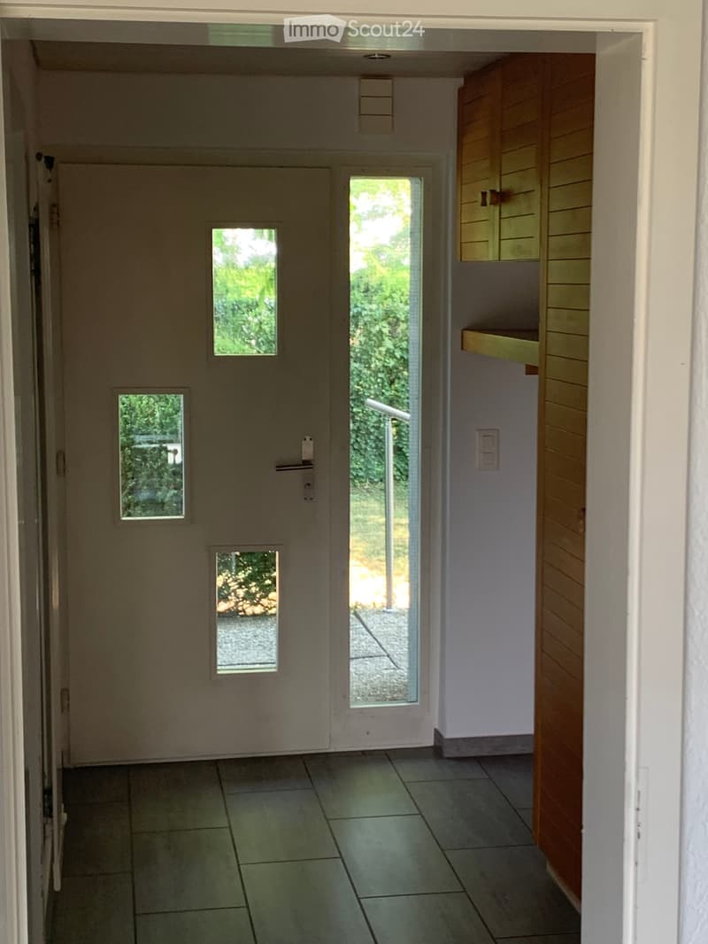 Empty hallway with tiled floor, white walls, and a wooden cabinet on the right side. A door with multiple small windows leads to an outdoor area.