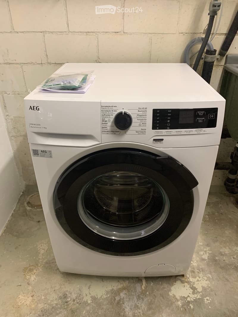 A white AEG washing machine with a control panel and a transparent lid, placed on a concrete floor next to a brick wall.