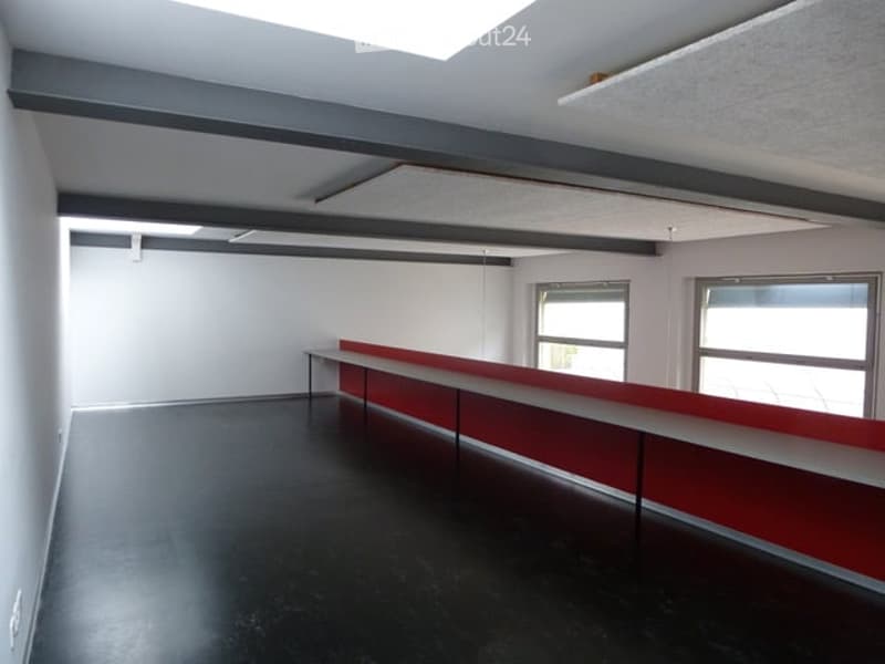 Empty room with white walls, black floor, and a red and white reception desk against one wall. Windows on the far side provide natural light.