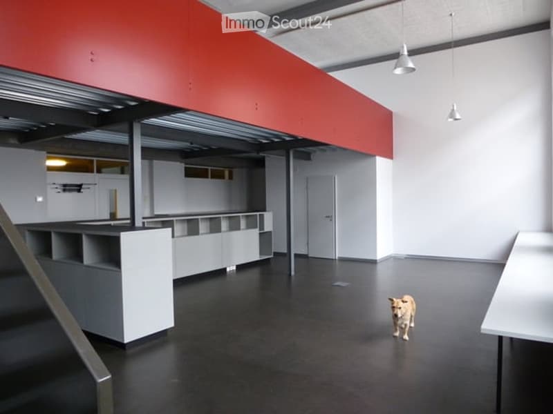 Modern, open-plan unfurnished room with white walls, black flooring, and a red accent wall. Features include built-in white shelving units, a grey staircase, and pendant lighting.