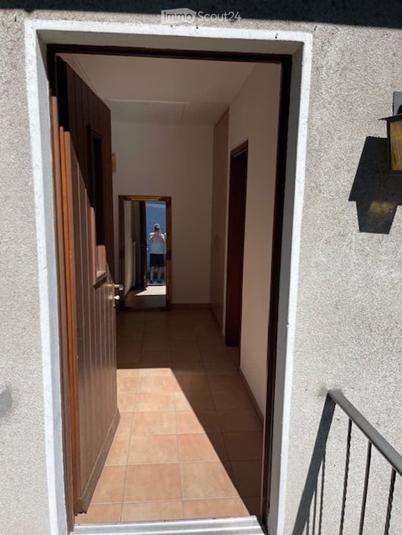 A narrow hallway with tiled flooring, white walls, and a wooden door on the left. A mirror is visible further down the hallway, and a metal railing is seen on the right side.
