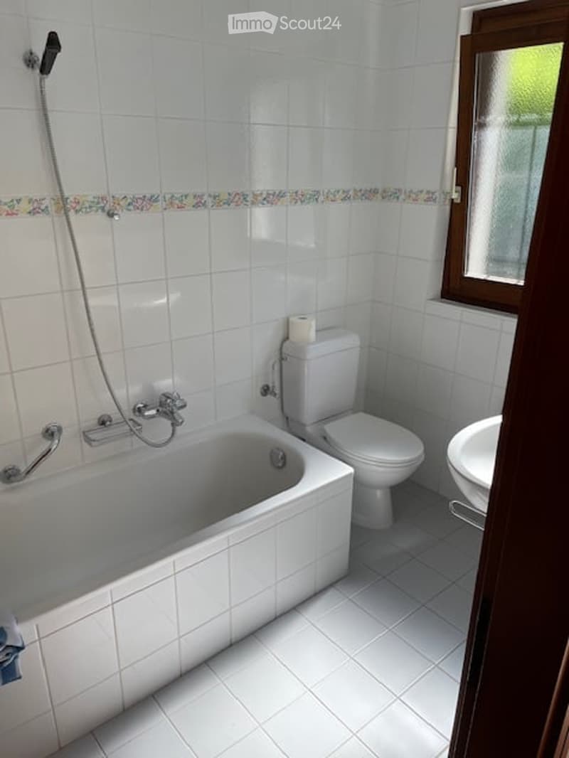 A small bathroom with white tiled walls and floor, featuring a bathtub, toilet, sink, and a shower hose attached to the wall.