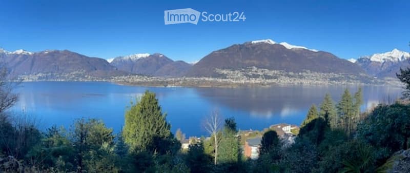 Panoramic view of a lake surrounded by snow-capped mountains, with trees and a small settlement visible on the shore.