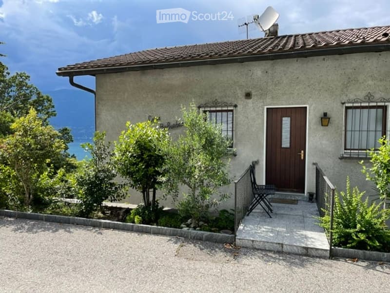 Single-story house with a tiled roof, satellite dish, and a small garden area with a bench. The house has a wooden front door and is surrounded by greenery with a scenic mountain view in the background.