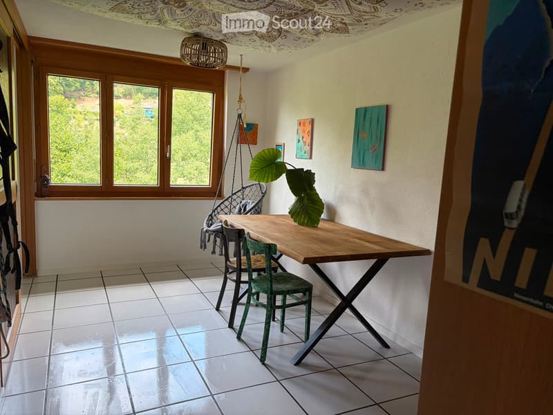 Bright room with large windows, white tiled floor, wooden table, two green chairs, wicker hanging chair, and a leafy plant on the table.