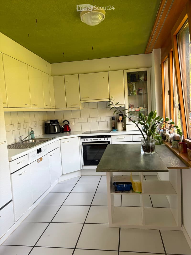 Bright kitchen with white tiled floors, light green upper cabinets, and a central island table. Appliances include an oven, dishwasher, and sink. A potted plant sits on the island, and various kitchen items are visible on the counters.