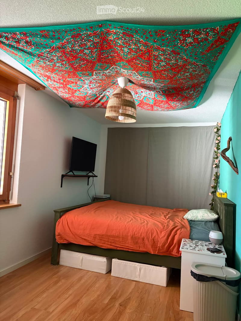 Cozy bedroom with a single bed, orange bedding, a nightstand with a sink, a wall-mounted TV, and a decorative tapestry on the slanted ceiling.