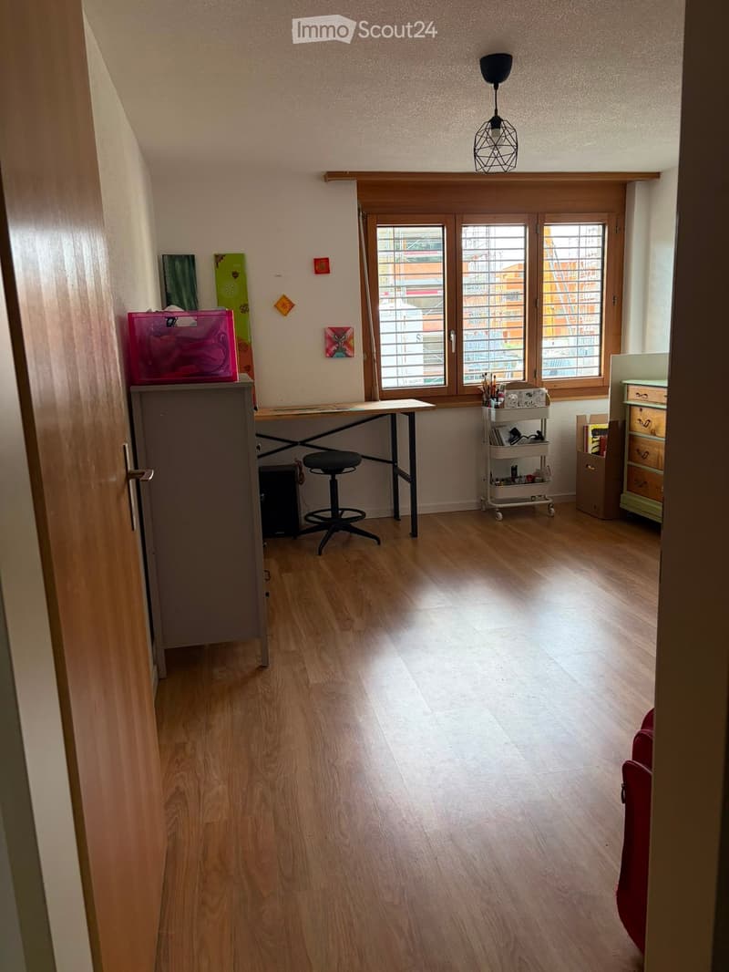 A well-lit room with wooden flooring, a desk, a stool, a small cart, and a cabinet. The room has large windows with blinds and a hanging light fixture.
