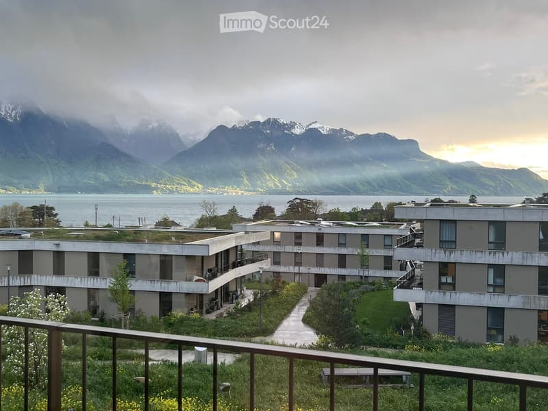 Modern multi-story building with balconies and a green roof, surrounded by a landscaped garden and overlooking a lake and mountains.