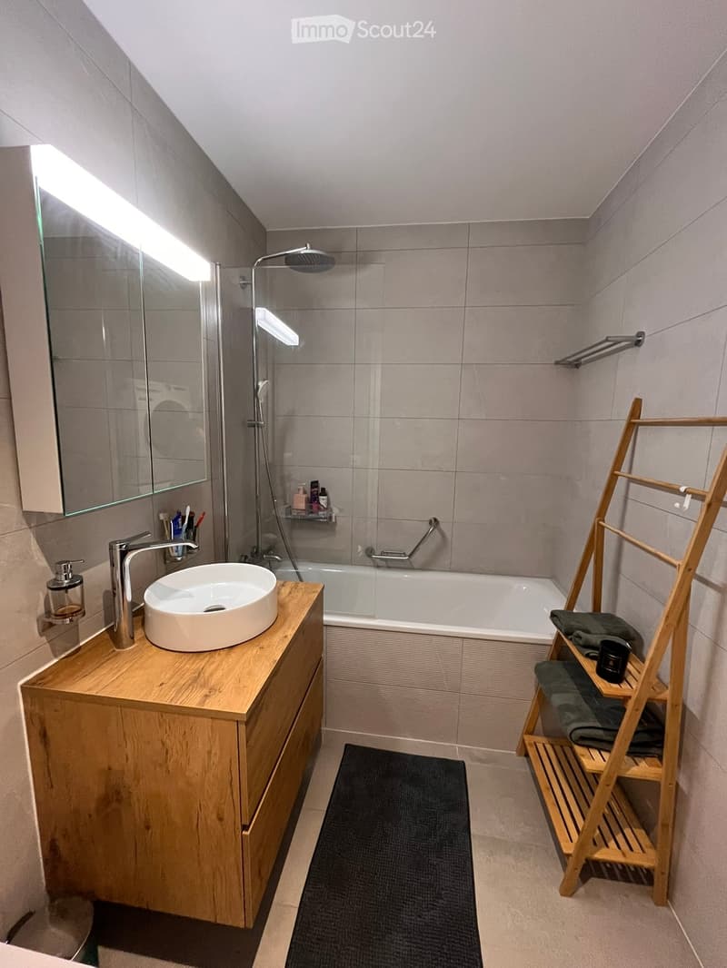 Modern bathroom with a wooden vanity, round sink, bathtub, mirror with built-in lighting, and a wooden towel rack with towels.