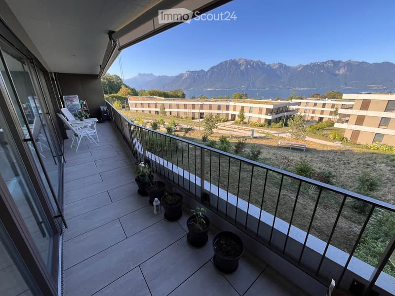 Balcony with tiled floor, glass railing, and scenic mountain and lake view. Includes white chairs, a small table, and several potted plants.