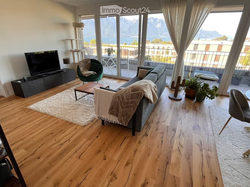Modern living room with wooden flooring, large windows offering a scenic mountain view, a grey sofa with a throw, a coffee table, a green armchair, a cat tree, a television, and potted plants.