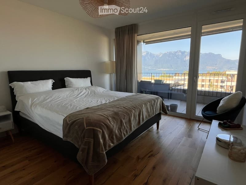 Modern bedroom with a large bed, nightstand, chair, and a view of mountains through large windows.