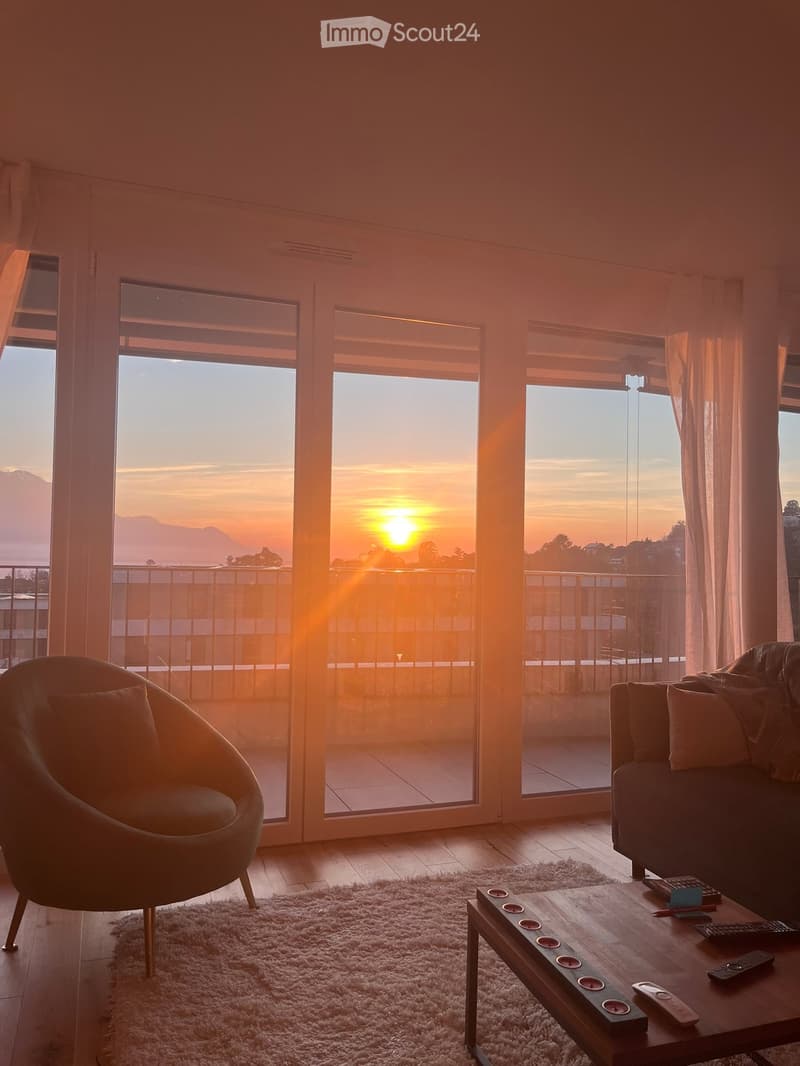 Living room with large windows offering a scenic sunset view, featuring a sofa, armchair, coffee table, and remote controls on the table.