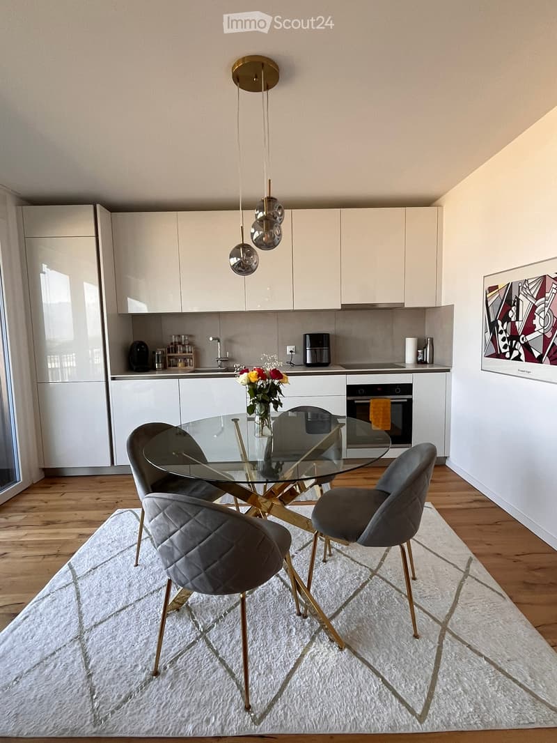 Modern kitchen and dining area with white cabinets, a glass dining table with four grey chairs, a white rug, and a vase of flowers on the table. The kitchen features stainless steel appliances and a sleek design.