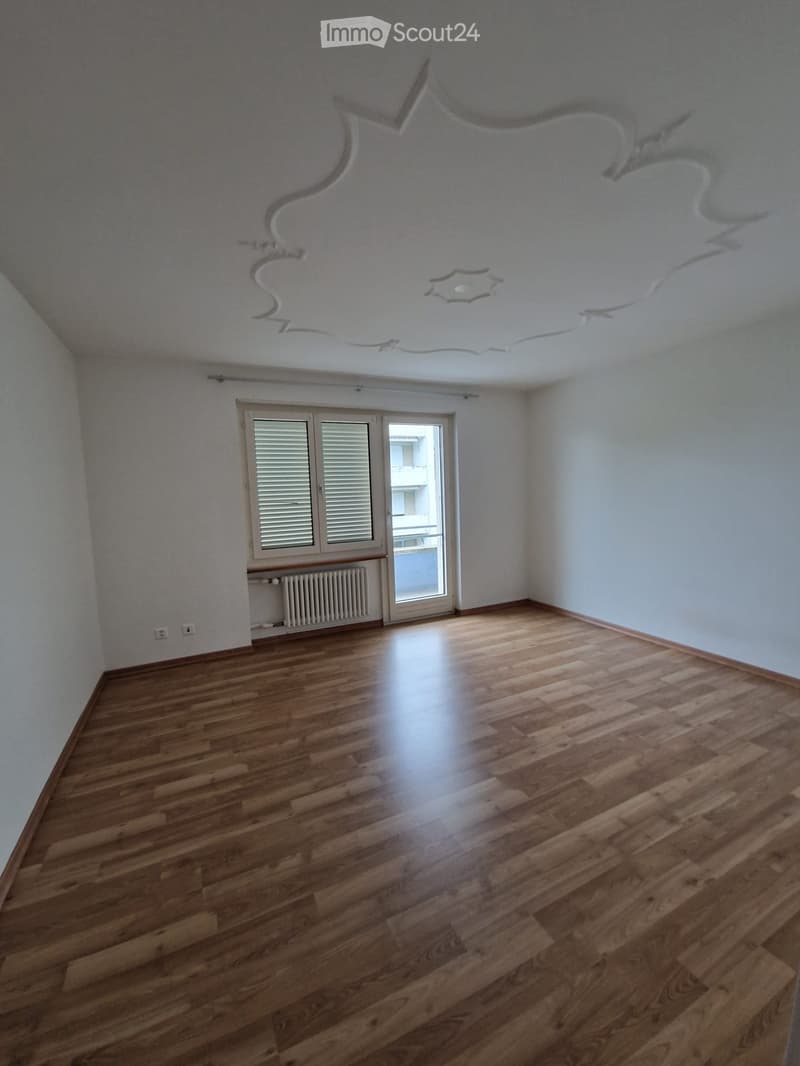 Empty room with wooden flooring, white walls, and a large window with blinds. The ceiling features a decorative design with a central light fixture.