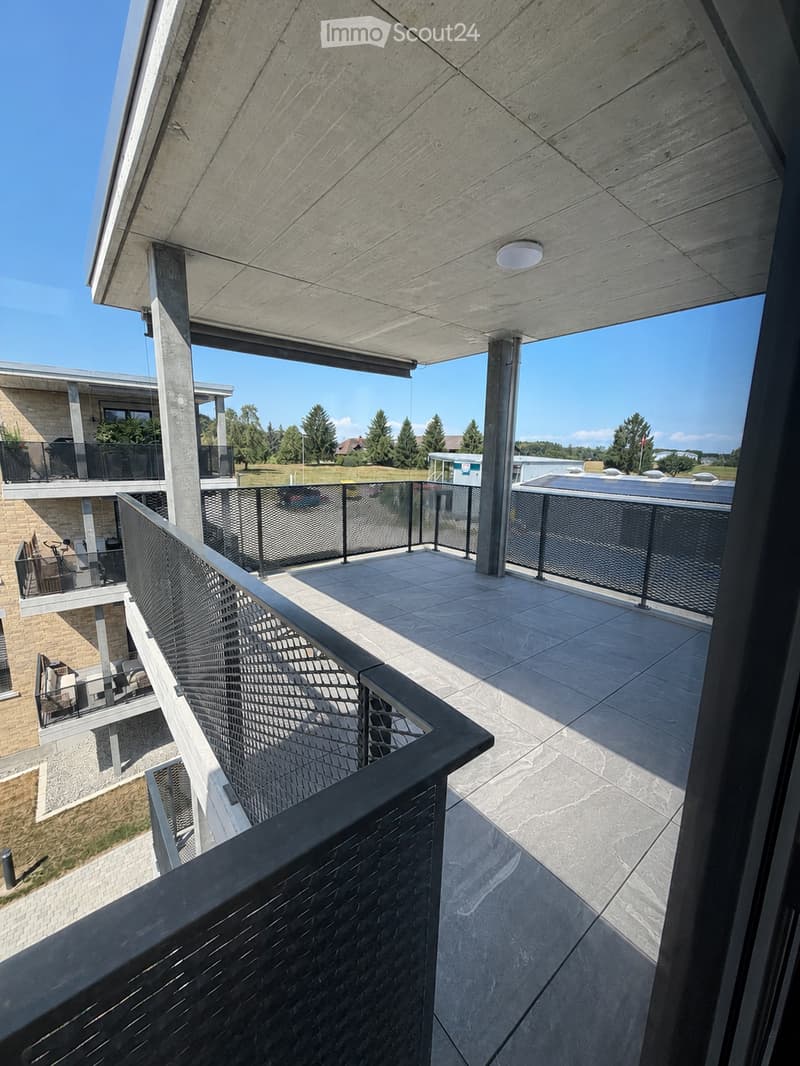 Modern balcony with stone tiles, black metal railing, and a covered section with a concrete roof. The view includes a grassy area, trees, and a parking lot.