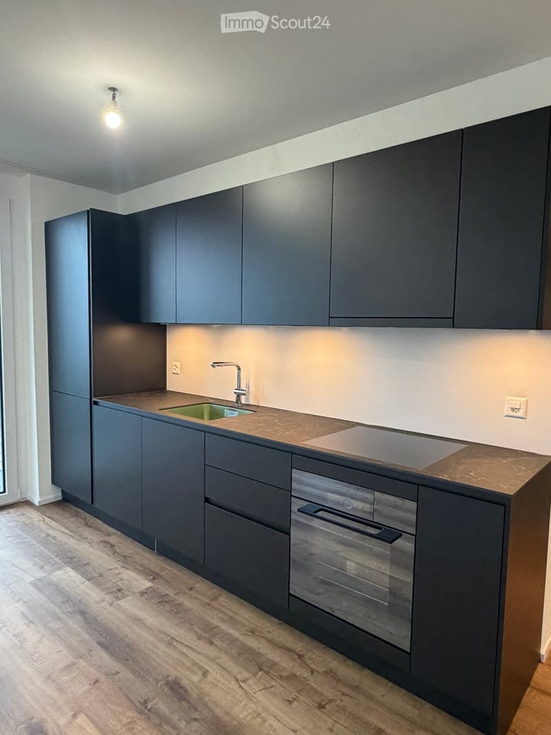 Modern kitchen with dark grey cabinets, a central island, a sink, and an oven. The countertops are a light brown material, and the floor is wooden.