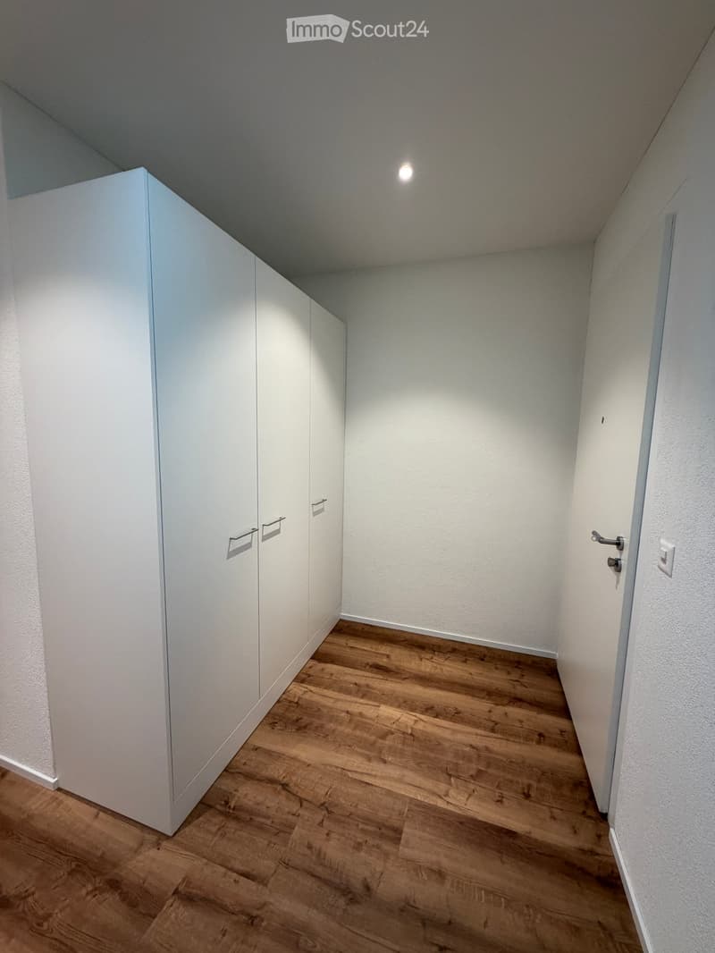 Empty hallway with wooden flooring, white walls, built-in white closet with silver handles, and a single light fixture on the ceiling.