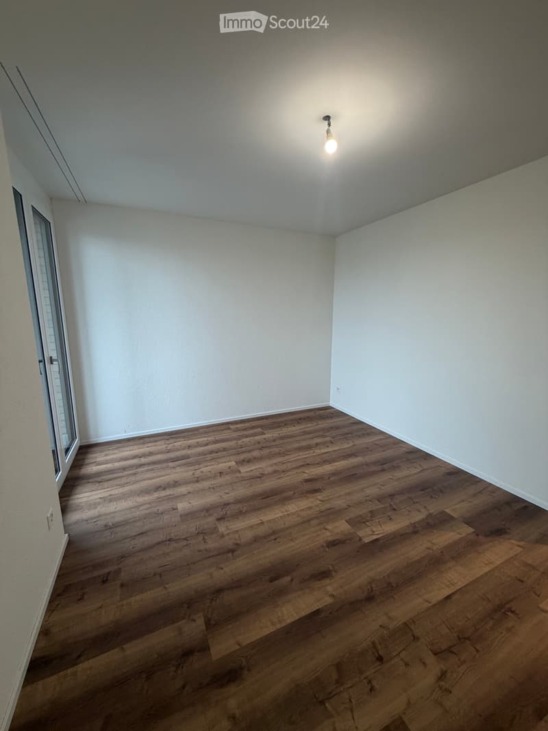 Empty room with wooden flooring and white walls, featuring a single ceiling light fixture and a window with a white frame.