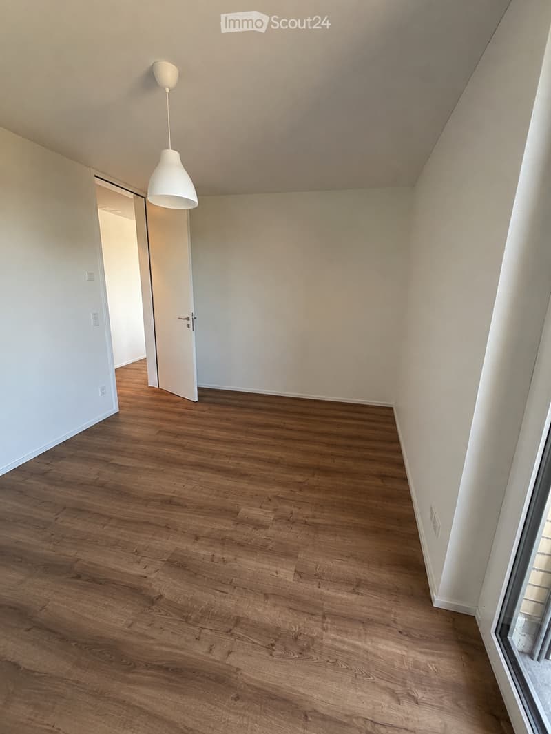 Empty room with wooden flooring, white walls, and a hanging light fixture. The room has a door leading to another space and a window on the right side.