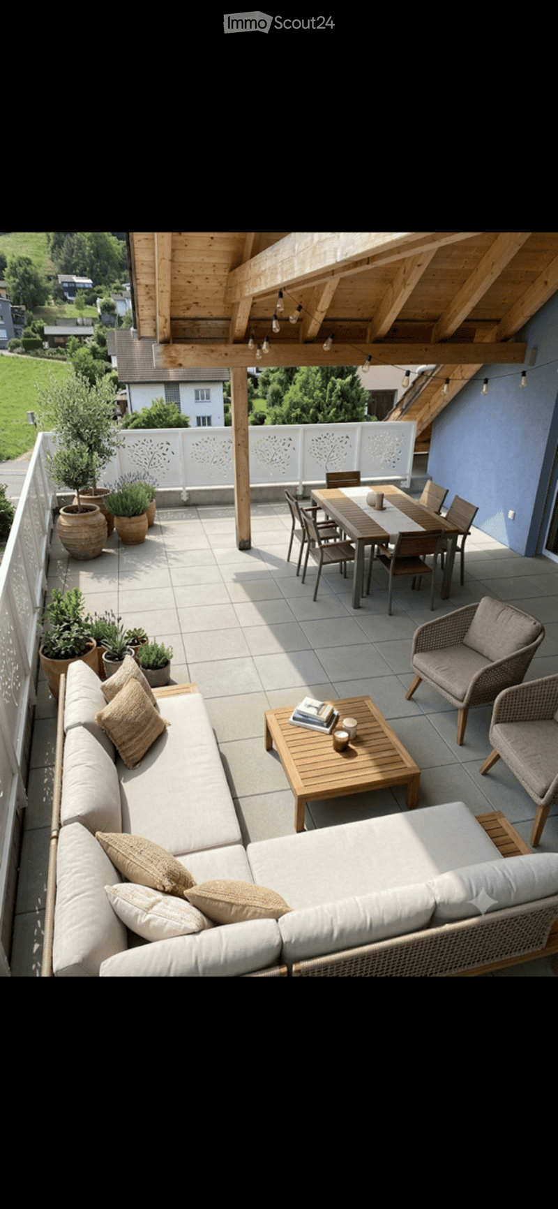 A spacious, well-lit balcony or terrace with a wooden pergola, outdoor seating including a sofa, coffee table, and dining set, surrounded by potted plants and offering a scenic view of a green landscape.