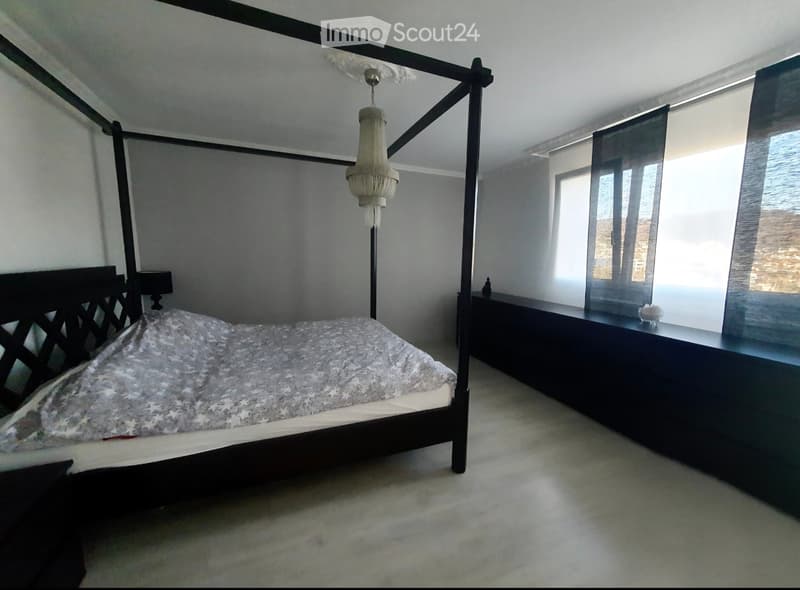 A modern bedroom with a large four-poster bed, a chandelier, and a nightstand with a lamp. The room has light-colored walls and carpeted flooring.