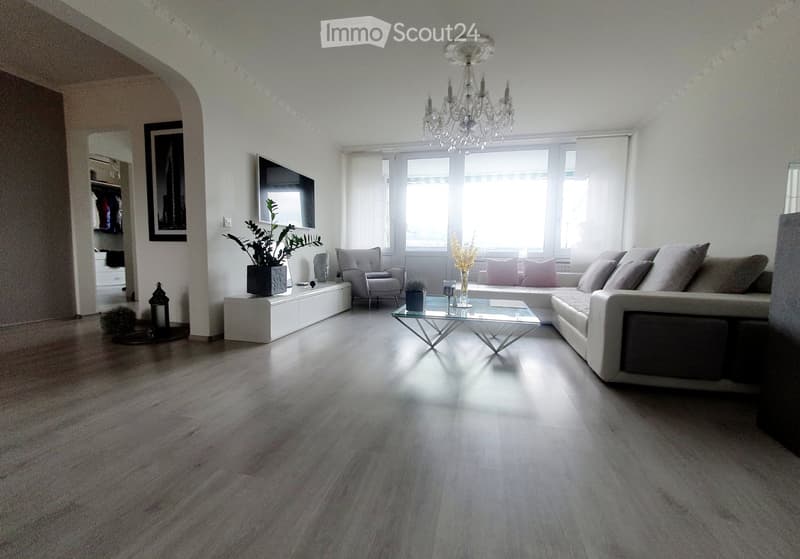Modern living room with light-colored wooden flooring, a large sectional sofa, a glass coffee table, an armchair, a chandelier, a TV, and decorative plants.