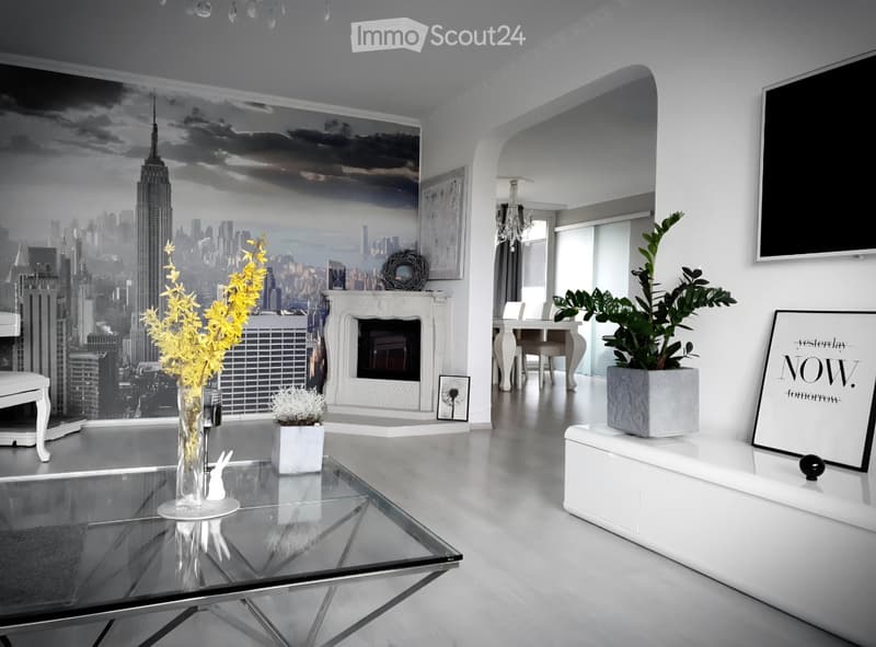 Modern living room with a glass coffee table, white sofa, fireplace, dining area, and decorative plants.