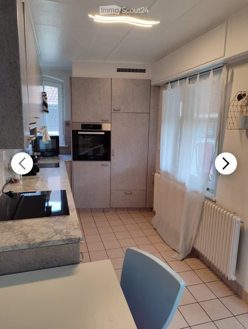 Modern kitchen with tiled floor, white cabinets, built-in oven, microwave, sink, and a radiator. A single chair is placed near the counter.
