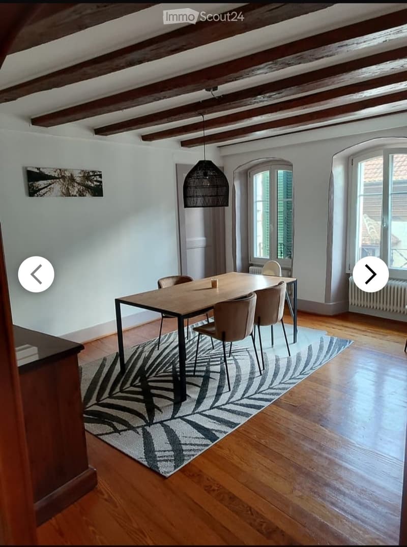 Dining room with wooden floor, exposed wooden beams, large windows, modern pendant light, dining table with chairs, and a patterned rug.