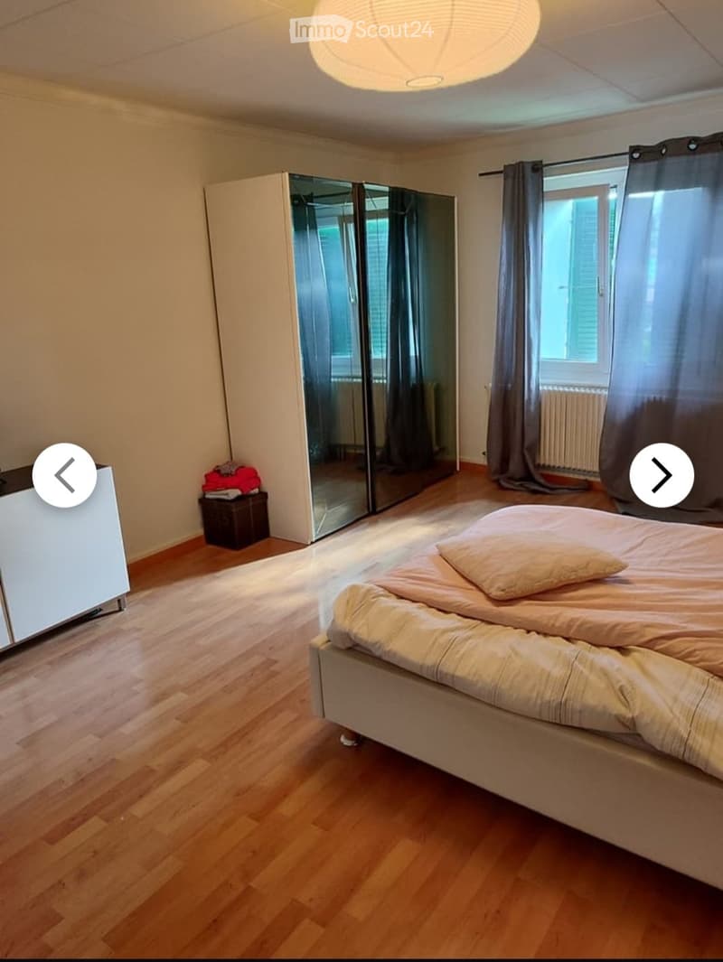 Bright bedroom with wooden flooring, a large bed with beige bedding, a mirrored wardrobe, and a window with curtains.