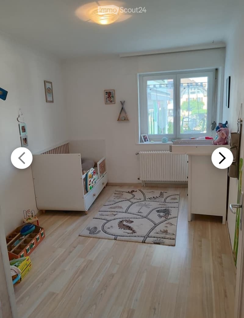 Brightly lit children's room with wooden flooring, a crib, a colorful rug, a toy box, and a radiator under a large window.