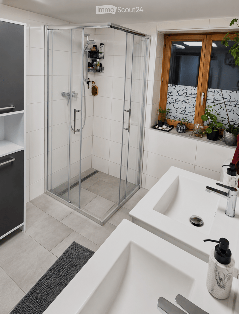 Modern bathroom with white tiles, a large shower enclosure, double sink vanity, bathtub, and decorative plants on the windowsill.