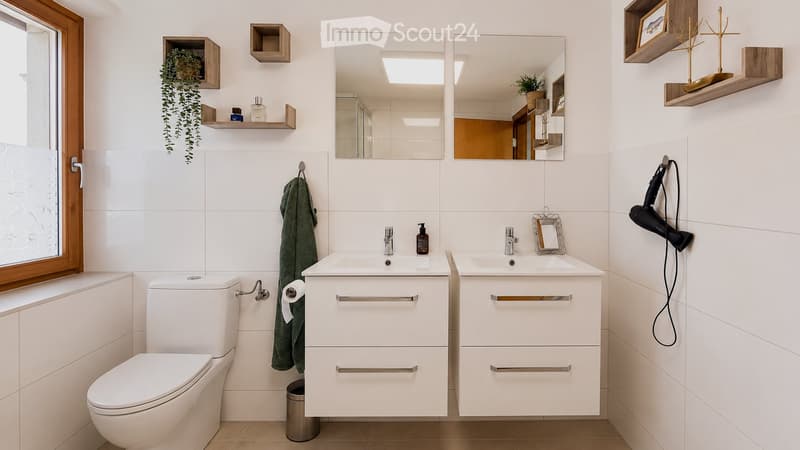 Modern bathroom with white tiles, double sink vanity, toilet, large mirror, towel, hair dryer, and decorative shelves with plants and picture frames.