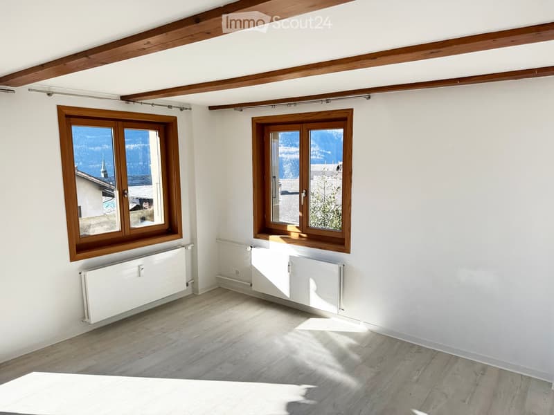 Empty room with wooden flooring, white walls, exposed wooden beams on the ceiling, and two large windows with wooden frames offering a view of the mountains.