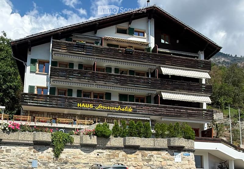 A multi-story wooden building with balconies, green shutters, and a stone base, surrounded by greenery and a mountainous backdrop.